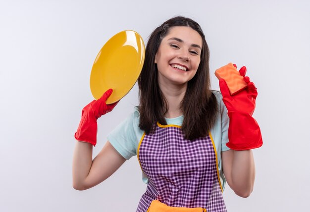 Hermosa joven en delantal y guantes de goma con plato y esponja sonriendo alegremente