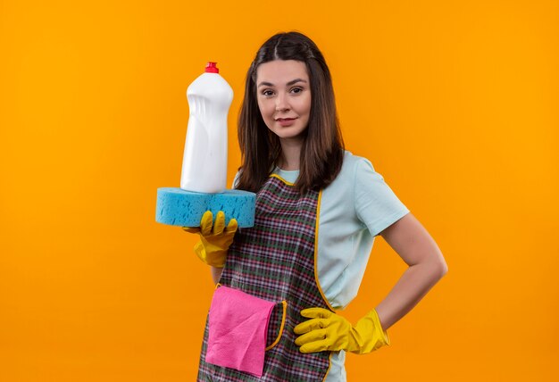 Hermosa joven en delantal y guantes de goma con productos de limpieza y esponja mirando a la cámara sonriendo confiado