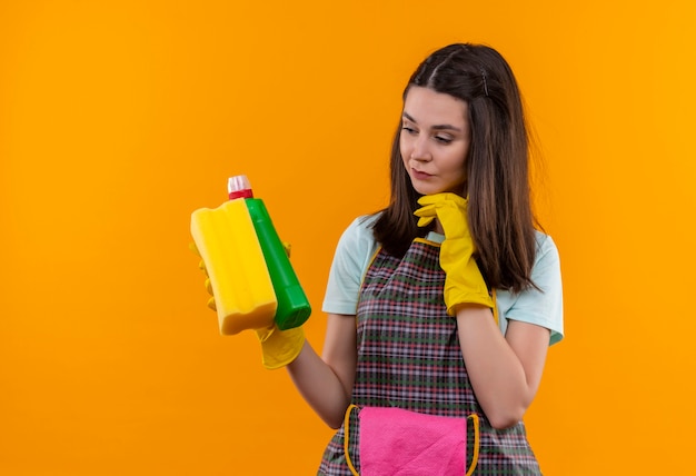 Foto gratuita hermosa joven en delantal y guantes de goma con productos de limpieza y una esponja mirándolos con expresión pensativa, tratando de tomar una decisión