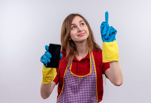 Hermosa joven en delantal y guantes de goma con smartphone apuntando con el dedo índice sonriendo