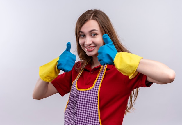 Hermosa joven en delantal y guantes de goma sonriendo mostrando los pulgares para arriba con ambas manos