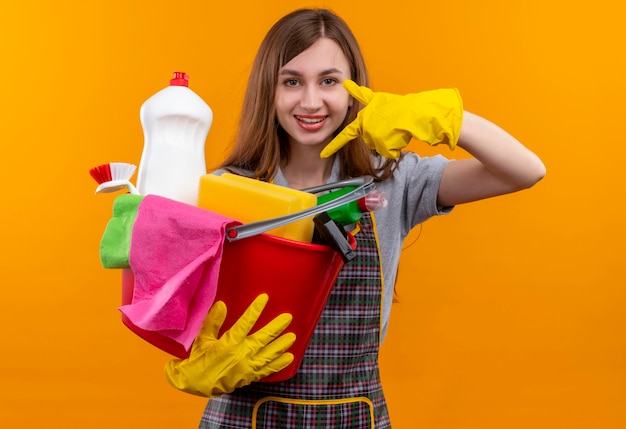 Hermosa joven en delantal y guantes de goma sosteniendo un balde con herramientas de limpieza apuntando con el dedo sonriendo alegremente