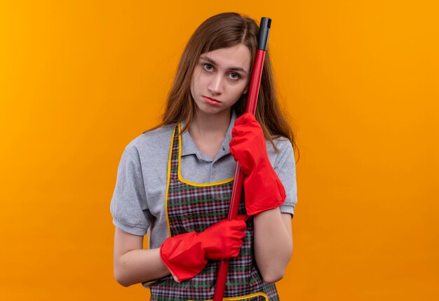 Hermosa joven en delantal y guantes de goma sosteniendo un trapeador con la cabeza inclinada sobre ella mirando a la cámara con expresión triste en la cara