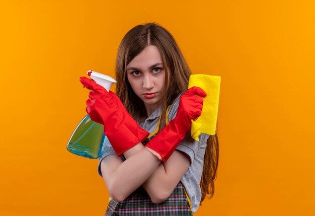 Hermosa joven en delantal y guantes de goma con spray y alfombra cruzando las manos haciendo gesto de defensa con cara seria