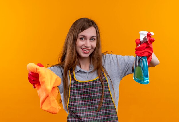 Hermosa joven en delantal y guantes de goma con spray de limpieza y alfombra mirando a la cámara sonriendo, listo para limpiar