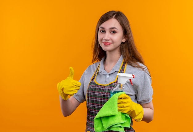 Hermosa joven en delantal y guantes de goma con spray de limpieza y alfombra mirando a la cámara sonriendo mostrando los pulgares para arriba, listo para limpiar