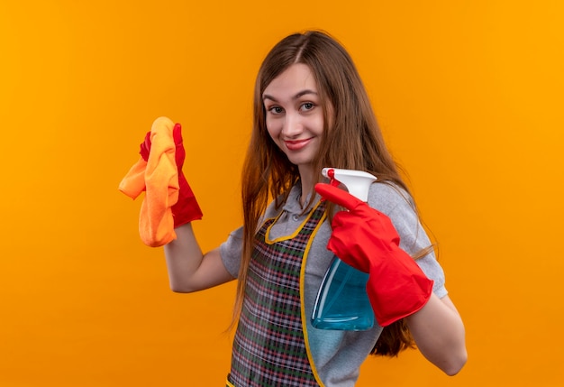 Hermosa joven en delantal y guantes de goma con spray de limpieza y alfombra mirando a la cámara sonriendo positivo y feliz, listo para limpiar