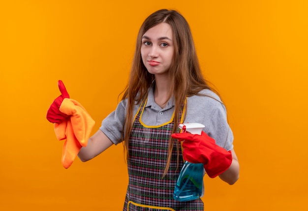 Hermosa joven en delantal y guantes de goma con spray de limpieza y alfombra mirando a la cámara con una sonrisa escéptica en la cara, mostrando los pulgares para arriba