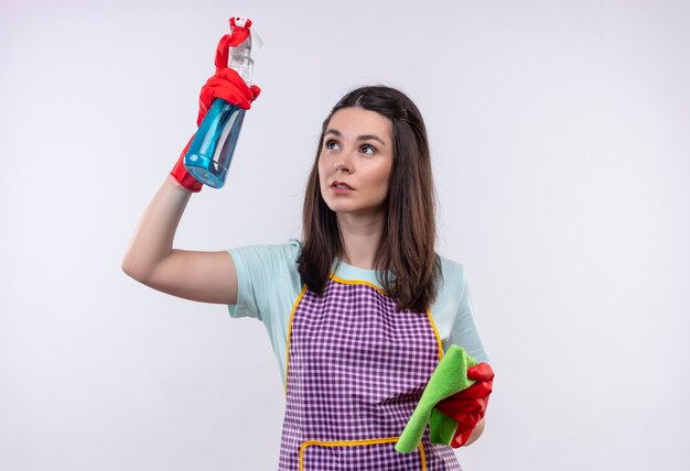 Hermosa joven en delantal y guantes de goma con spray de limpieza y alfombra que va a limpiar mirando confiado