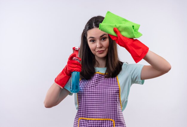 Hermosa joven en delantal y guantes de goma con spray de limpieza y alfombra sonriendo confiado, listo para limpiar