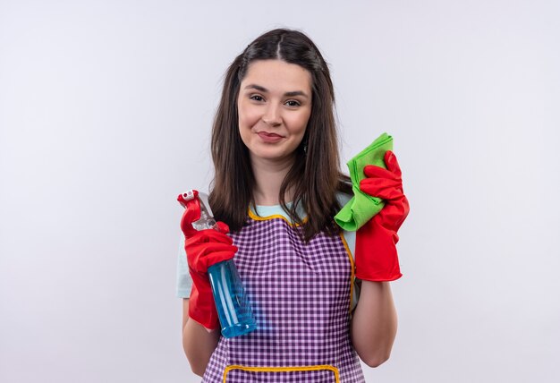 Hermosa joven en delantal y guantes de goma con spray de limpieza y alfombra sonriendo confiado, listo para limpiar