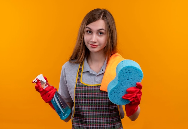 Hermosa joven en delantal y guantes de goma con spray de limpieza y esponja mirando a la cámara sonriendo, listo para limpiar