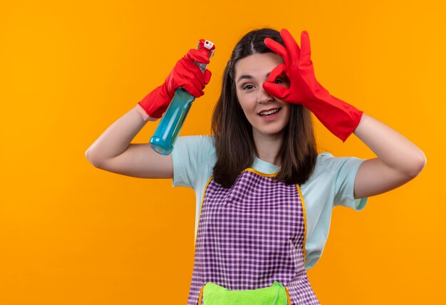 Hermosa joven en delantal y guantes de goma con spray de limpieza sonriendo alegremente haciendo bien firmar mirando a través de este signo