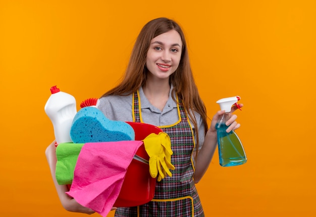 Hermosa joven en delantal sosteniendo balde con herramientas de limpieza y spray sonriendo alegremente