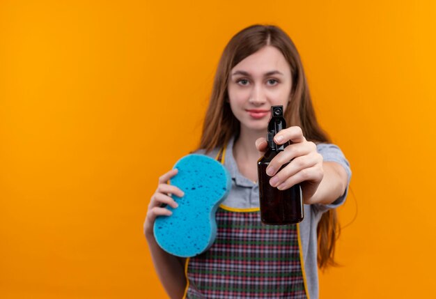 Hermosa joven en delantal sosteniendo spray limpiador y esponja mirando a la cámara sonriendo amable, listo para limpiar