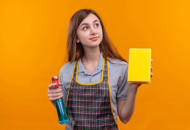 Hermosa joven en delantal sosteniendo spray limpiador y esponja mirando a un lado con mirada soñadora sonriendo