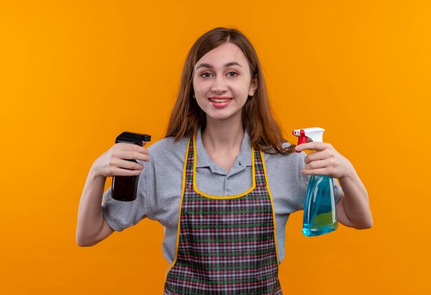 Hermosa joven en delantal sosteniendo suministros de limpieza mirando a la cámara sonriendo amable, listo para limpiar