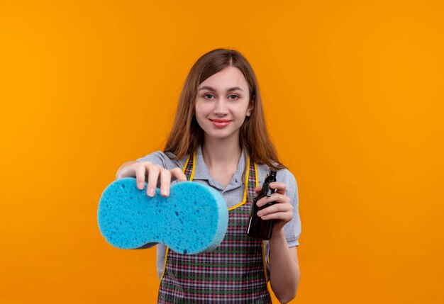 Hermosa joven en delantal con spray de limpieza y esponja mirando a la cámara sonriendo, listo para limpiar