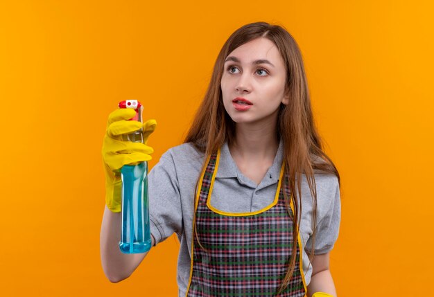 Hermosa joven en delantal con spray de limpieza mirando a un lado, yendo a limpiar