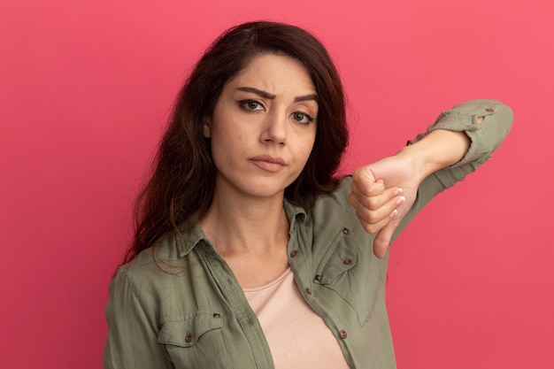 Foto gratuita hermosa joven disgustada con camiseta verde oliva que muestra el pulgar hacia abajo aislado en la pared rosa