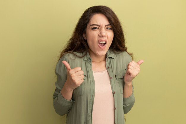 Hermosa joven disgustada con camiseta verde oliva que muestra los pulgares para arriba aislados en la pared verde oliva