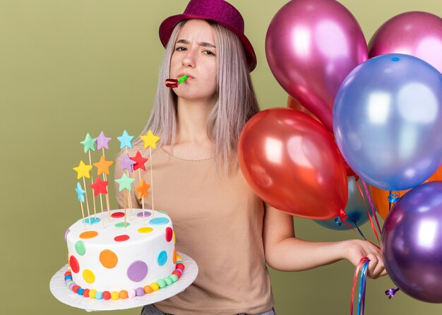Hermosa joven disgustada con gorro de fiesta sosteniendo globos con silbato de fiesta soplado de pastel