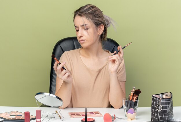 Hermosa joven disgustada sentada en el escritorio con herramientas de maquillaje sosteniendo y mirando pinceles de maquillaje aislado sobre fondo verde oliva