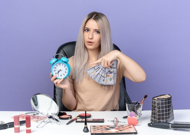 Hermosa joven disgustada se sienta a la mesa con herramientas de maquillaje con dinero en efectivo y puntos en el reloj de alarma en su mano aislado sobre fondo azul.