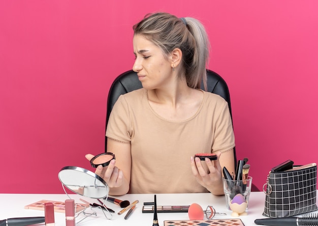 Hermosa joven disgustada se sienta a la mesa con herramientas de maquillaje con rubor en polvo aislado sobre fondo rosa