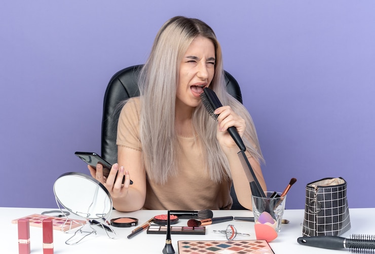 Foto gratuito hermosa joven disgustada se sienta a la mesa con herramientas de maquillaje sosteniendo un peine con teléfono aislado sobre fondo azul.