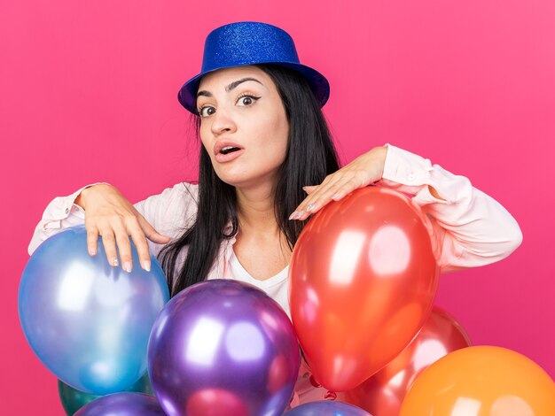 Hermosa joven disgustada con sombrero de fiesta de pie detrás de globos aislados en la pared rosa