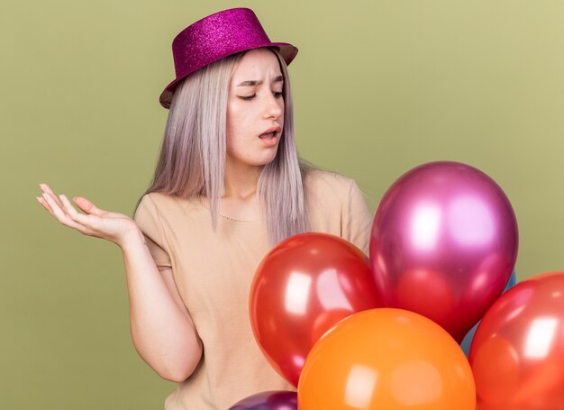 Hermosa joven disgustada con sombrero de fiesta de pie detrás de globos extendiendo la mano aislada en la pared verde oliva