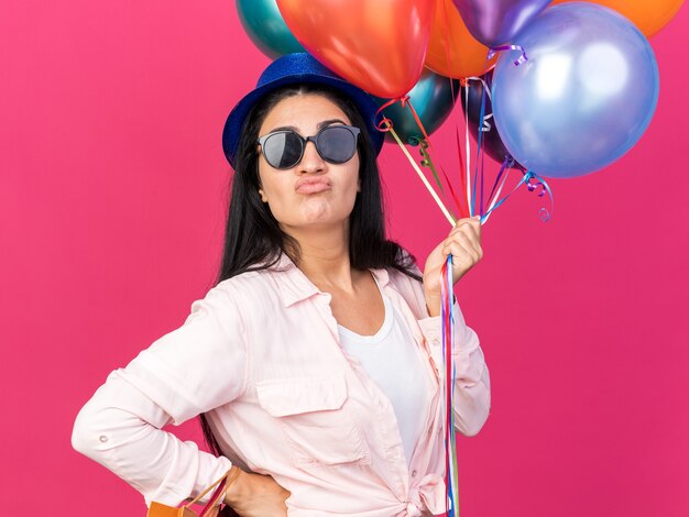 Hermosa joven disgustada con sombrero de fiesta sosteniendo globos con bolsas de regalo poniendo la mano en la cadera aislada en la pared rosa