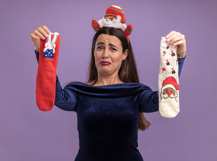 Foto gratuito hermosa joven disgustada con vestido azul y aro de pelo de navidad sosteniendo calcetines de navidad en cámara aislada sobre fondo púrpura