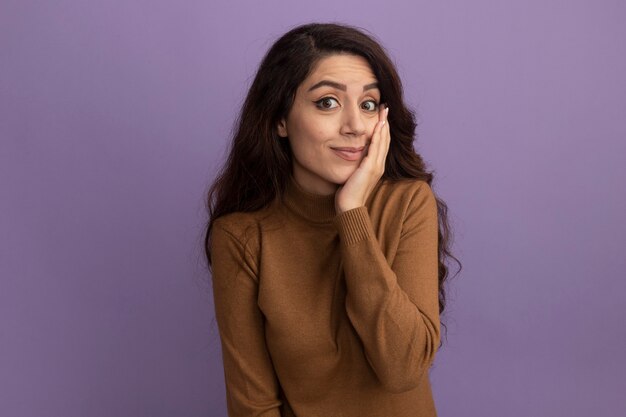 Hermosa joven disgustada vistiendo un suéter de cuello alto marrón poniendo la mano en la mejilla aislada en la pared púrpura