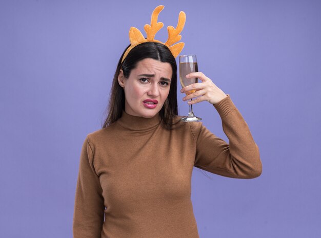 Hermosa joven disgustada vistiendo suéter marrón con aro de pelo de Navidad sosteniendo una copa de champán en el hombro aislado sobre fondo azul.