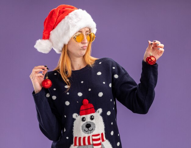 Hermosa joven disgustada vistiendo un suéter de Navidad y sombrero con gafas sosteniendo y mirando bolas de Navidad aisladas sobre fondo púrpura