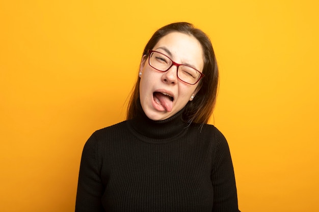 Hermosa joven divertida en un cuello alto negro guiñando un ojo sacando la lengua feliz y positivo