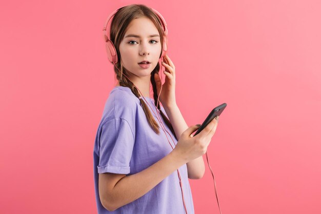 Hermosa joven con dos trenzas en camiseta lila con auriculares sosteniendo el celular en la mano mientras mira soñadoramente en cámara sobre fondo rosa