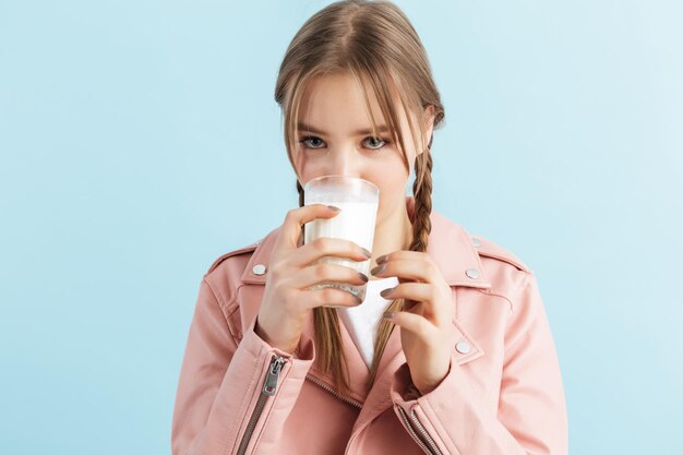 Hermosa joven con dos trenzas en chaqueta de cuero rosa y camiseta blanca bebiendo leche mientras mira soñadoramente en cámara sobre fondo azul.