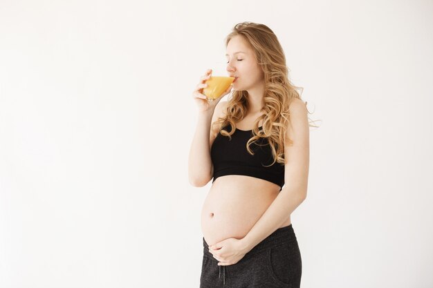 Hermosa joven embarazada de pelo claro en traje negro cómodo vaso de jugo de naranja con los ojos cerrados y expresión relajada