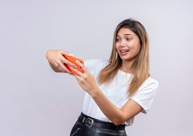 Una hermosa joven emocionada en camiseta blanca jugando en el teléfono móvil