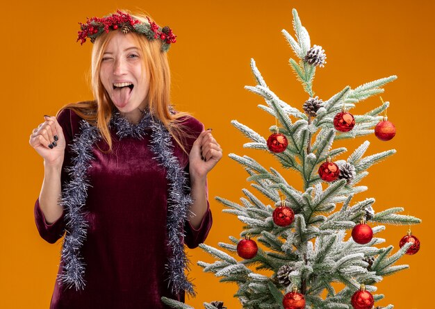 Hermosa joven emocionada de pie cerca del árbol de Navidad con vestido rojo y corona con guirnalda en el cuello mostrando la lengua y haciendo gesto de sí aislado en la pared naranja