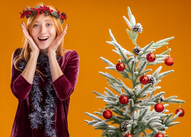 Hermosa joven emocionada de pie cerca del árbol de Navidad con vestido rojo y corona con guirnalda en el cuello tomados de la mano alrededor de la cara aislada en la pared naranja