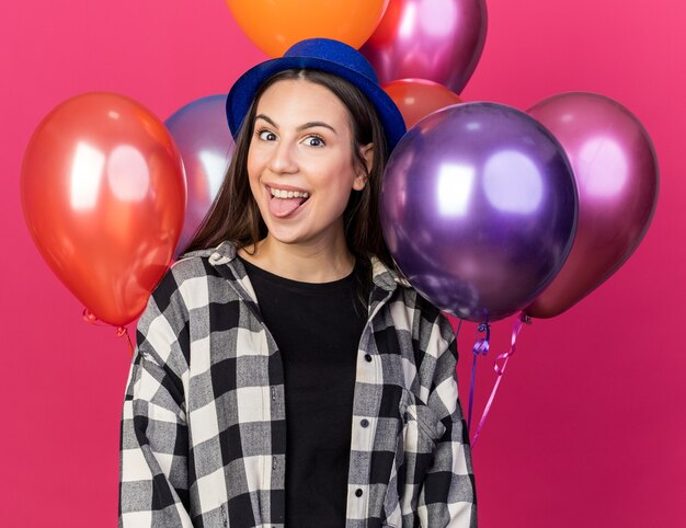 Hermosa joven emocionada con sombrero de fiesta de pie delante de globos mostrando lengua aislada en pared rosa