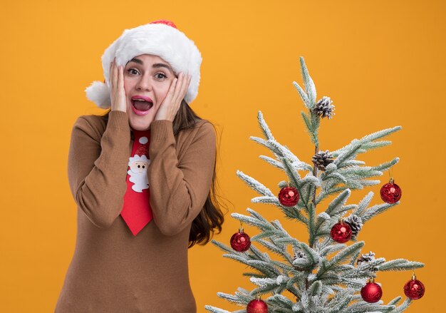 Hermosa joven emocionada con sombrero de navidad con corbata de pie cerca del árbol de navidad poniendo las manos en las mejillas aisladas en la pared naranja