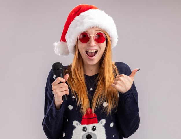 Hermosa joven emocionada con sombrero de navidad y gafas sosteniendo el micrófono mostrando el pulgar hacia arriba aislado sobre fondo blanco.