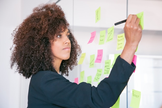 Hermosa joven empresaria escribiendo en etiqueta con marcador. gerente femenina rizada profesional concentrada compartiendo idea para proyecto y tomando nota