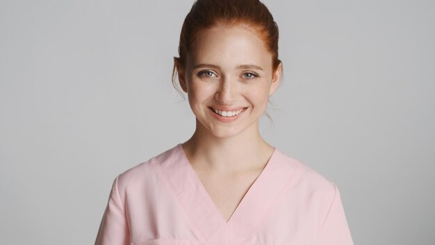 Hermosa joven enfermera sonriente en uniforme felizmente mirando en cámara sobre fondo blanco.