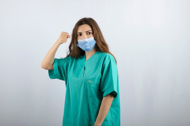 Hermosa joven enfermera en uniforme verde con una máscara médica y mostrando un puño